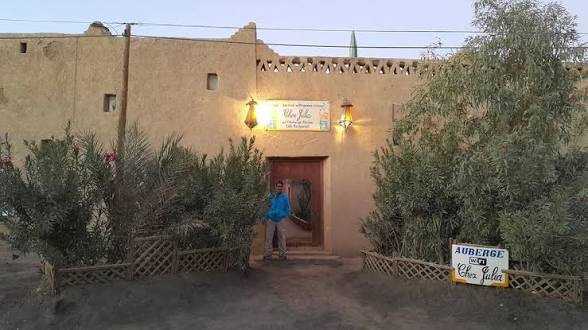 Auberge building with beige adobe walls, wooden door, greenery at entrance, lit sign reading Chez Julia supporting Camel Trekking in the Sahara Desert tours.