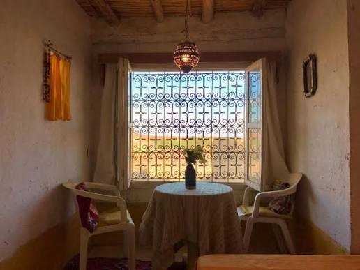 Cozy rustic Moroccan room with white textured walls, a round table with a lace cloth, two plastic chairs, and a decorative window grill, capturing the ambiance of Camel Trekking in the Sahara Desert.