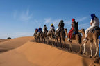 Camel trekking with riders wearing colorful headscarves crossing golden sand dunes under a clear blue sky