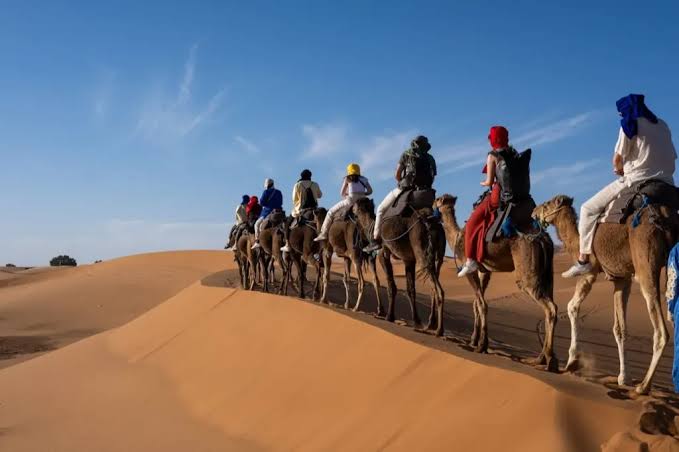 Camel trekking with riders wearing colorful headscarves crossing golden sand dunes under a clear blue sky