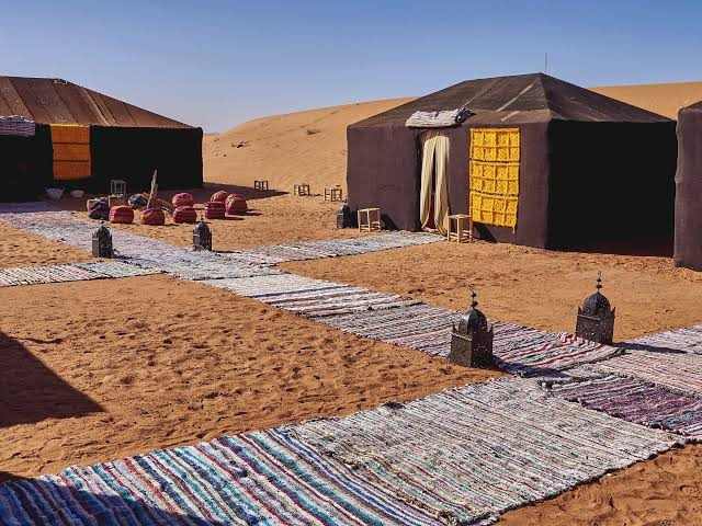 Traditional black desert tents with yellow accents set on sand with patterned rugs and lanterns for Camel Trekking in Erg Chebbi