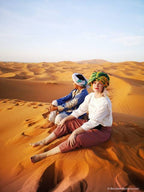 Camel Trekking experience showing two people in traditional colorful Moroccan headwear and desert clothing seated on golden sand dunes of Erg Chebbi under clear sky