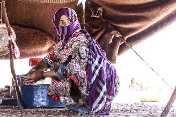 Woman wearing colorful patterned dress and a purple checkered scarf sitting under a makeshift tent, preparing for camel trekking