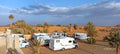 Campground les Roches Merzouga with white recreational vehicles and palm trees on sandy desert terrain under a blue sky with clouds.