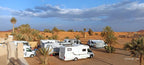 Campground les Roches Merzouga with white recreational vehicles and palm trees on sandy desert terrain under a blue sky with clouds.