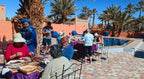 Outdoor dining area at Campground les Roches Merzouga with people seated around tables covered in purple cloths near a swimming pool and palm trees.