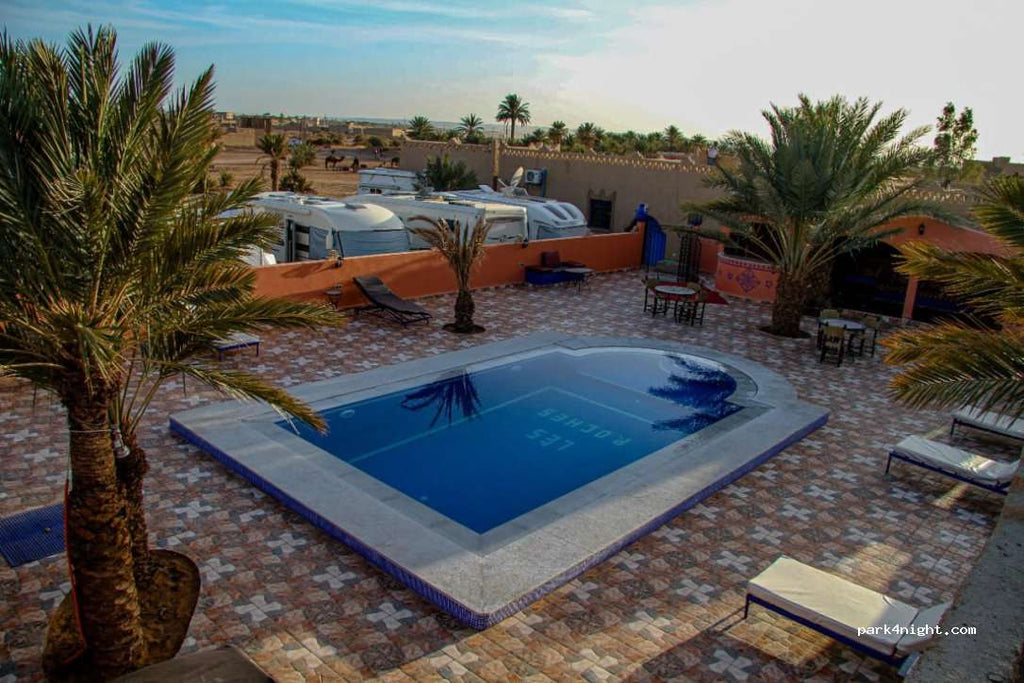 Campground les Roches Merzouga outdoor area with rectangular blue swimming pool, tiled floor, palm trees, and lounge chairs around.