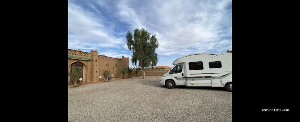 Campground les Roches Merzouga with a light brown desert-style building, gravel ground, green trees, and a white motorhome parked nearby under a partly cloudy sky.