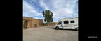 Campground les Roches Merzouga with a light brown desert-style building, gravel ground, green trees, and a white motorhome parked nearby under a partly cloudy sky.