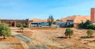 Camping La Gazelle Bleue features white desert tents with blue trim, set on sandy terrain near orange sand dunes and sparse greenery.