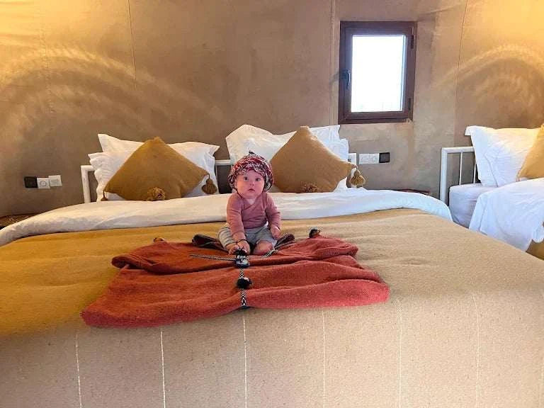 Camping La Gazelle Bleue room with beige and white bedding, mustard cushions, and a baby sitting on a rust-colored blanket.