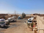 Camping La Gazelle Bleue with white motorhomes parked on a dry desert ground beside traditional brown mud walls and sparse trees under a clear blue sky