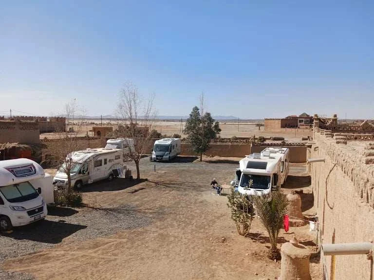 Camping La Gazelle Bleue with white motorhomes parked on a dry desert ground beside traditional brown mud walls and sparse trees under a clear blue sky
