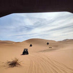 Camping La Gazelle Bleue showing sandy desert dunes with three all-terrain vehicles riding under a partly cloudy sky.