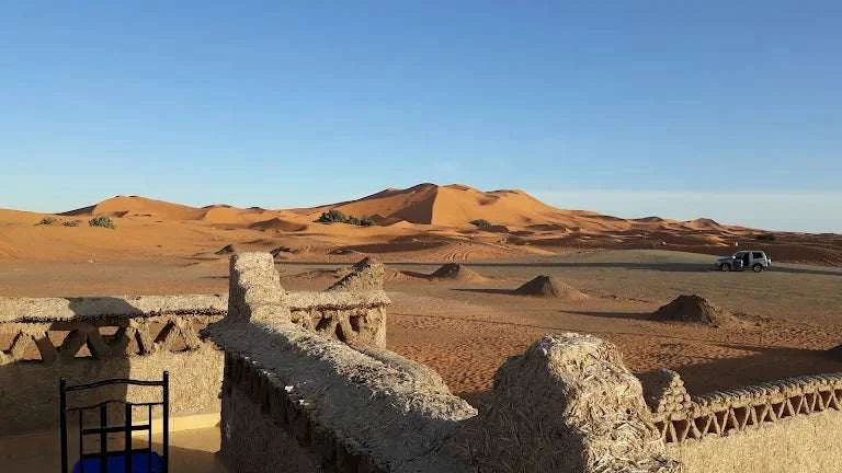 Desert camp structure with textured beige walls and a blue chair overlooking orange sand dunes under a clear sky at Camping La Gazelle Bleue