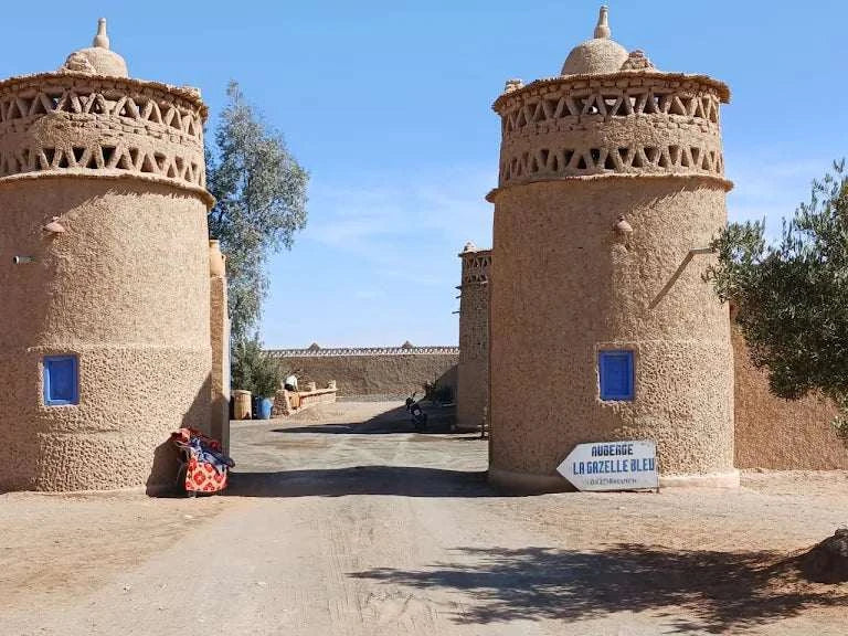 Entrance gate towers of Camping La Gazelle Bleue made of textured beige adobe with decorative geometric top patterns and small blue windows under clear sky