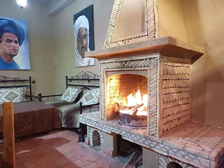 Rustic beige tiled fireplace with burning fire inside, beside metal-framed beds with patterned cushions in a traditional Moroccan room at Camping La Gazelle Bleue.