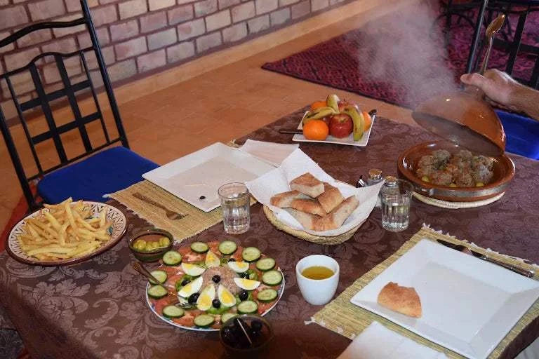 Dining table with Moroccan meal featuring a round vegetable and egg salad, steaming tagine with meatballs, fresh bread, fries, and fruit plate at Camping La Gazelle Bleue