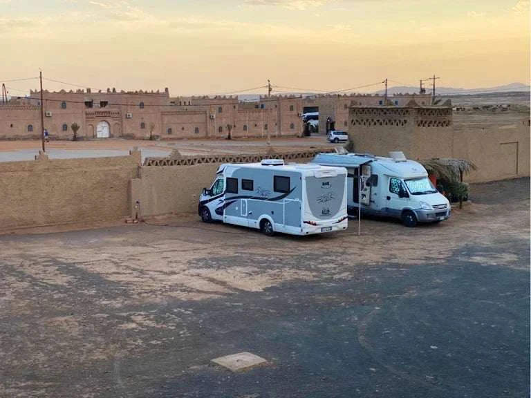 Camping La Gazelle Bleue showing two white and light gray motorhomes parked next to a beige adobe wall in a desert setting at dusk.
