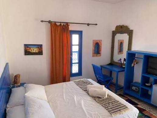 Chez Youssef Guesthouse bedroom with white walls, blue wooden furniture, orange curtain, and neatly made bed with white linens