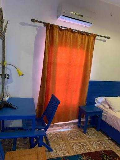 Chez Youssef Guesthouse room with blue wooden bed, matching desk and chair, orange curtain, white walls, and air conditioning