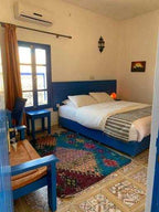 Chez Youssef Guesthouse bedroom with blue wooden bed frame, patterned rug, tiled floor, and beige walls with window and rustic lamp