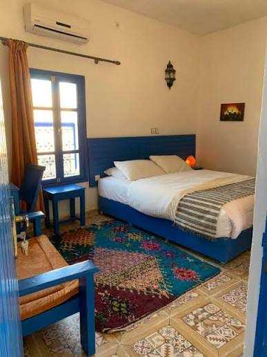 Chez Youssef Guesthouse bedroom with blue wooden bed frame, patterned rug, tiled floor, and beige walls with window and rustic lamp