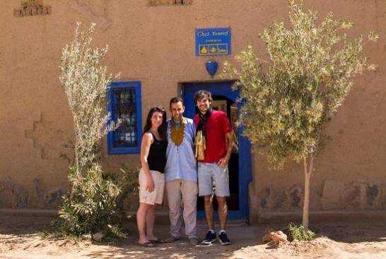 Chez Youssef Guesthouse exterior showing three people standing in front of a traditional beige stucco building with blue windows and door, two small trees nearby