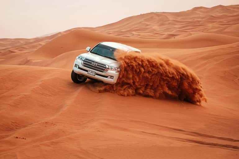 White SUV kicking up red sand while driving on large desert dunes during Day Tour from Erfoud to Merzouga adventure.