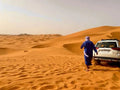 4x4 vehicle parked on golden sand dunes with a person in traditional blue attire walking beside it during a Day Tour from Erfoud to Merzouga in the Sahara desert.