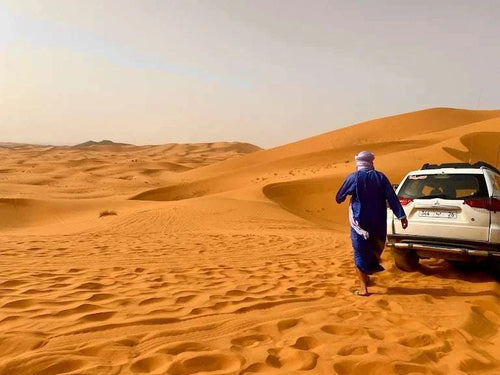 4x4 vehicle parked on golden sand dunes with a person in traditional blue attire walking beside it during a Day Tour from Erfoud to Merzouga in the Sahara desert.