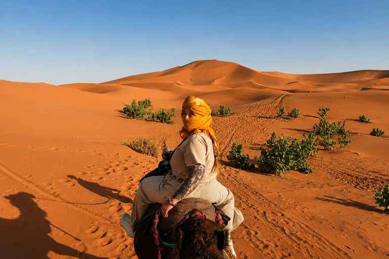 Woman wearing beige clothing and a yellow headscarf riding a camel across golden sand dunes and sparse green bushes during Day Tour from Erfoud to Merzouga