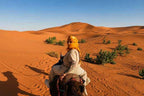 Woman wearing beige clothing and a yellow headscarf riding a camel across golden sand dunes and sparse green bushes during Day Tour from Erfoud to Merzouga