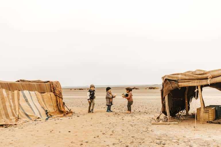 Desert camp tents with striped fabric and three children standing on sandy terrain during a Day Tour from Erfoud to Merzouga in the Sahara Desert.