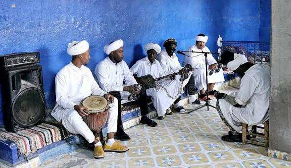 Group of men dressed in traditional white robes and turbans playing drums and string instruments in a blue room during Day Tour from Erfoud to Merzouga