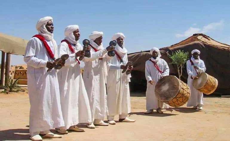 Group of six men dressed in traditional white robes and turbans playing hand drums and percussion outdoors during Day Tour from Erfoud to Merzouga.
