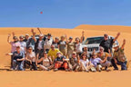 Group of people in casual desert attire posing on golden sand dunes with two white 4x4 vehicles under clear blue sky during a Day Tour from Erfoud to Merzouga