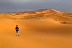 Desert landscape with golden sand dunes under gray sky and a person walking, illustrating a Day Tour from Erfoud to Merzouga.