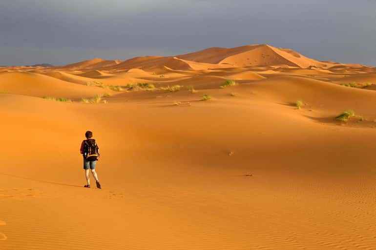Desert landscape with golden sand dunes under gray sky and a person walking, illustrating a Day Tour from Erfoud to Merzouga.
