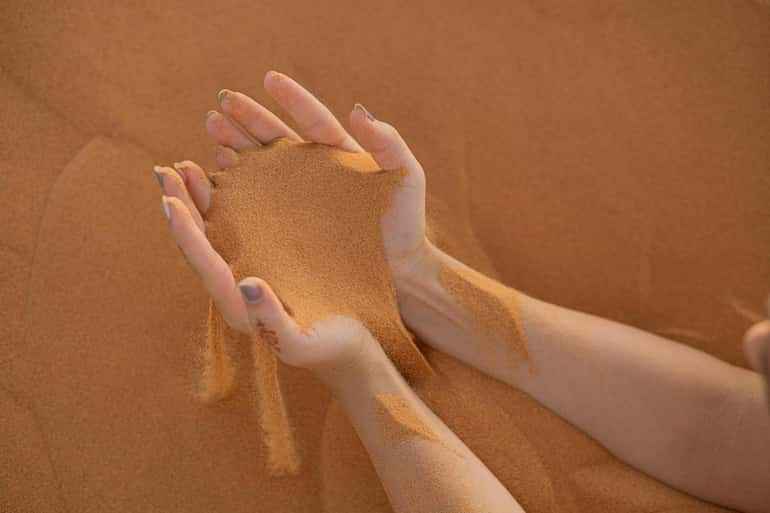 Hands holding and letting flow fine golden desert sand, creating a soft stream against a sandy background for Day Tour from Erfoud to Merzouga.