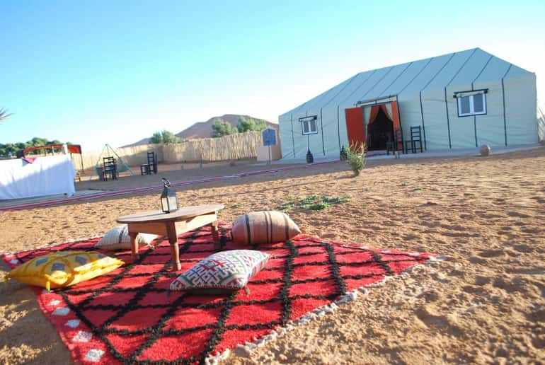 Moroccan desert camp with beige canvas tents, red patterned rug, low wooden table, and colorful cushions on sand, ideal for Day Tour from Erfoud to Merzouga.