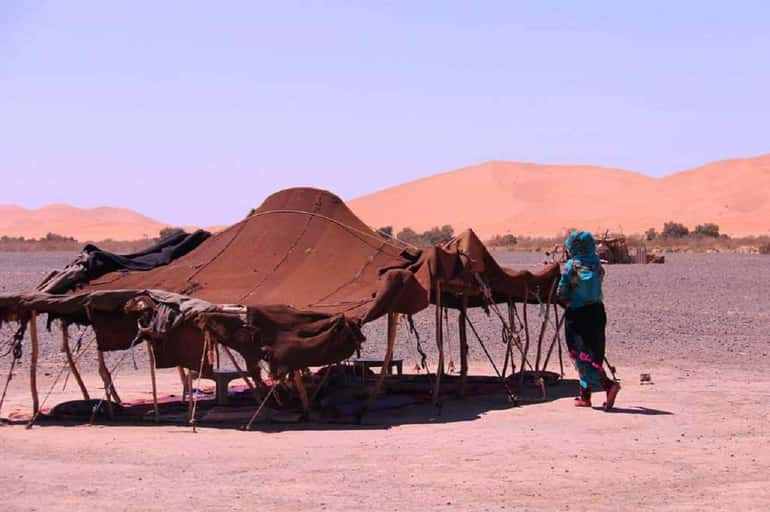 Traditional brown desert tent made of fabric and leather with a person in blue attire standing next to it in a sandy landscape, highlighting a Day Tour from Erfoud to Merzouga.