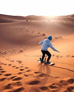 Person wearing a grey hoodie and blue hat sandboarding on golden dunes at sunset during a Day Tour from Erfoud to Merzouga