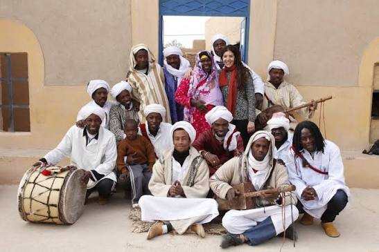 Group of traditionally dressed musicians and a tourist posing together outside a beige building, showcasing cultural heritage on a Day Tour from Erfoud to Merzouga.