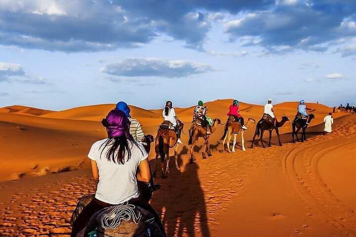 Day Tour from Fes to Merzouga showing group camel trekking with riders in colorful headscarves across orange sand dunes under blue sky
