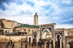 Day Tour from Fes to Merzouga showing historic beige stone buildings and ornate archways with green-tiled roofs under a cloudy sky