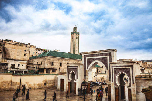 Day Tour from Fes to Merzouga showing historic beige stone buildings and ornate archways with green-tiled roofs under a cloudy sky