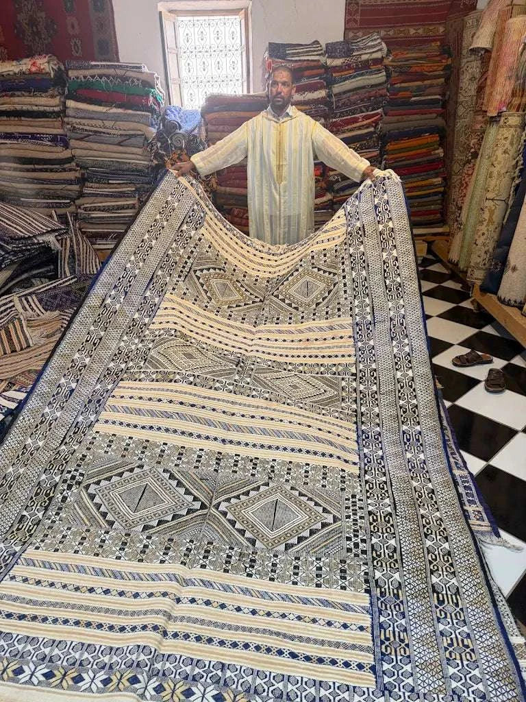 Moroccan man holding large beige and black patterned rug with geometric designs in a textile shop at Dépôt Nomade