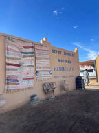 Exterior view of Dépôt Nomade with beige wall displaying colorful striped woven rugs and traditional Moroccan handicrafts.