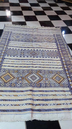 Moroccan woven rug with geometric patterns in blue, brown, and white colors displayed on a checkered floor at Dépôt Nomade.